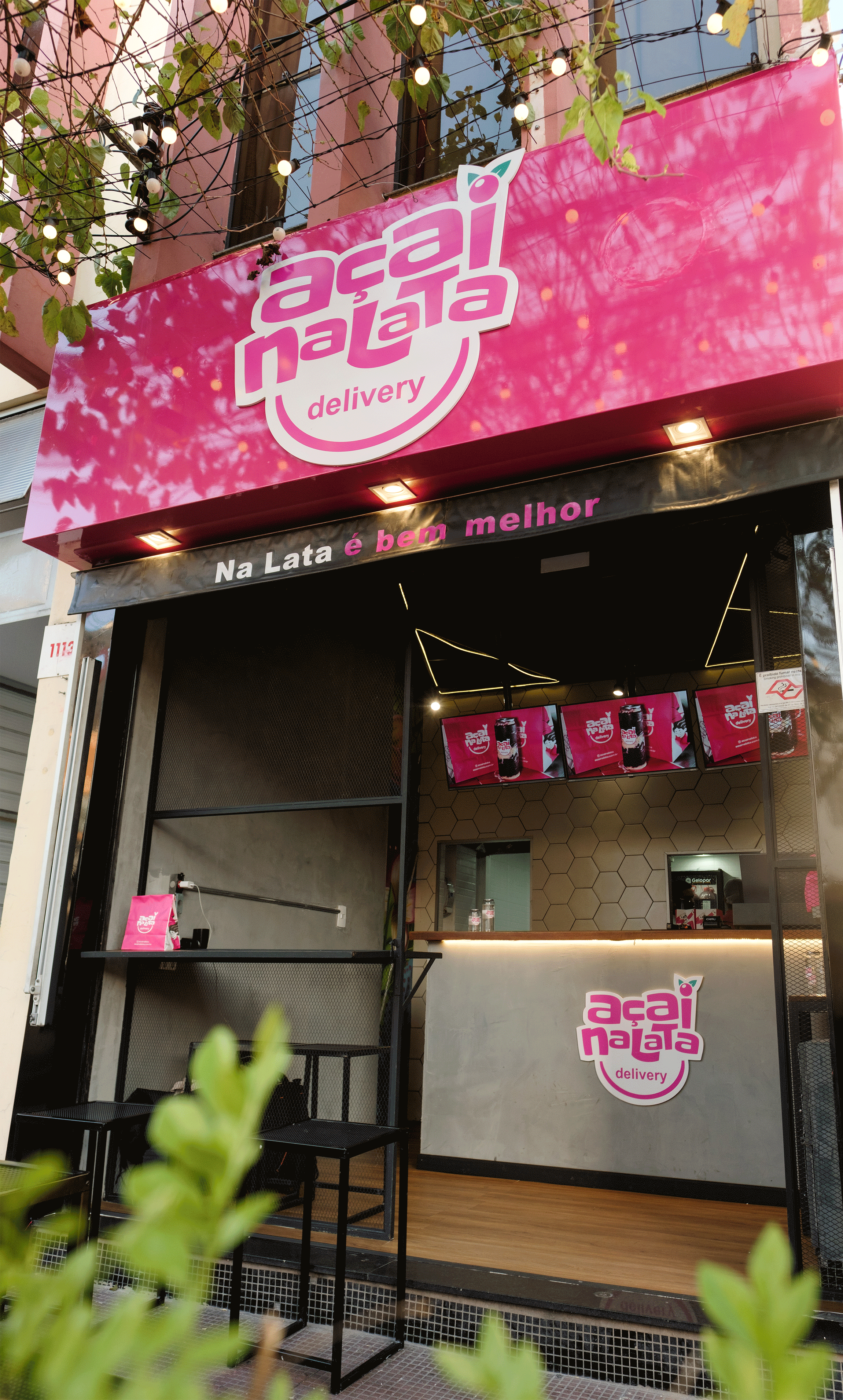 Fachada de uma loja Açaí NaLata durante o dia, com letreiro rosa iluminado, varal de lâmpadas e o balcão de atendimento ao fundo